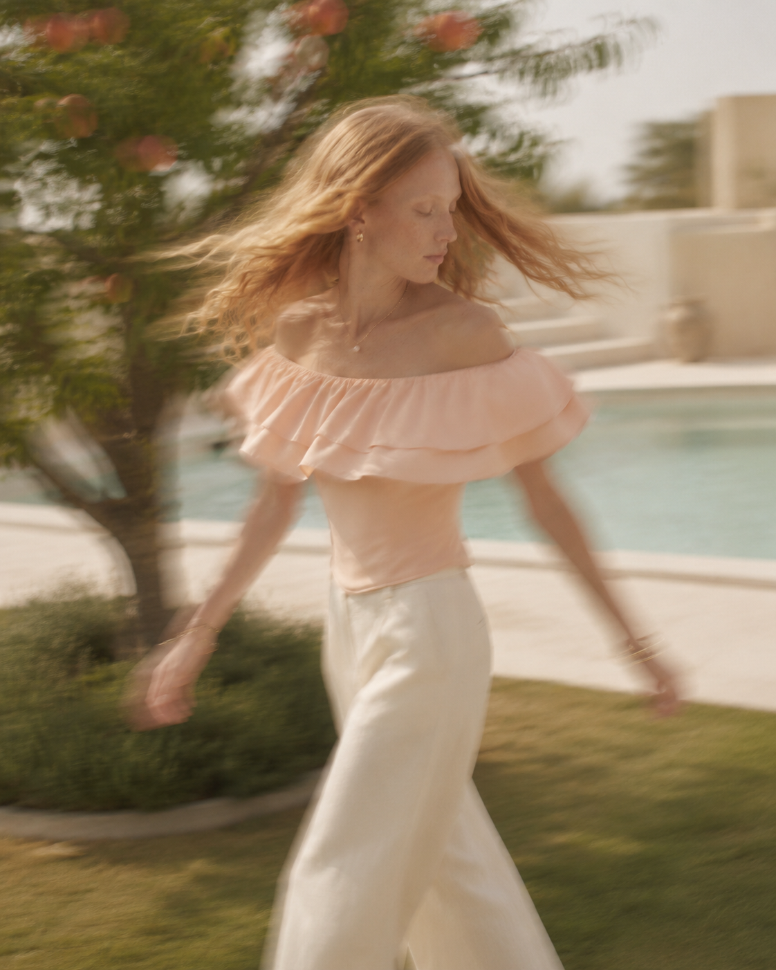 VKELLER Ruth peach off-shoulder ruffle silk top, with white trousers, walking by the pool.